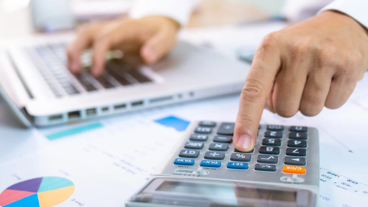 person with one hand typing on a laptop and the other working a calculator